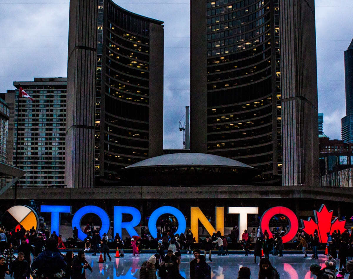 Toronto sign in front of buildings downtown at dusk with people outside Toronto sign in front of buildings downtown at dusk with people outside